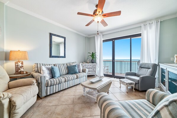 A spacious living room with a large window, a ceiling fan, and a view of the ocean. - BEACHFRONT, FAMILY FRIENDLY, LOW FLOOR, INDOOR/OUTDOOR POOL | BEACHBALL PROPERTIES (Orange Beach)