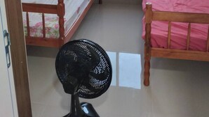 A small room with two wooden beds, one with a floral bedspread and the other with a pink bedspread. There is a window with a view of buildings and trees outside. A black fan is on the floor.