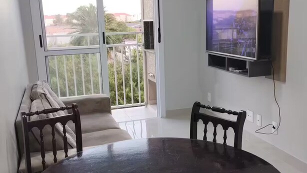 A dining area with a round table and chairs, a TV mounted on the wall, and a sliding glass door leading to a balcony with a view of the outside.