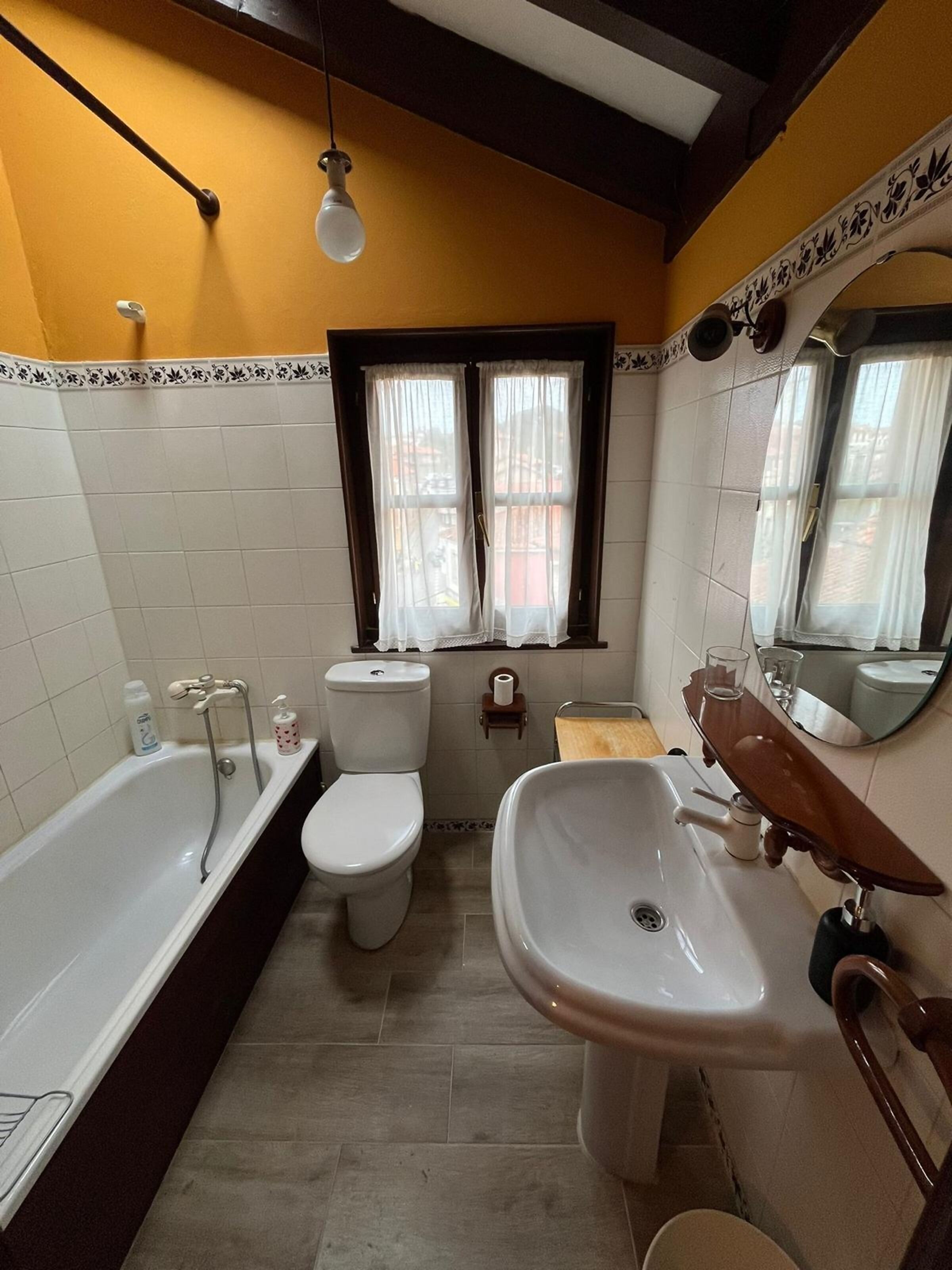 A bathroom with a bathtub, toilet, and sink, featuring a window with curtains, a towel rack, and a mirror.