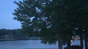 A wooden deck with a metal railing overlooking a body of water, with trees and a distant building visible. - Portage Lakes, Lakefront, Private Dock & Deck (Akron)