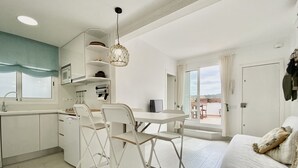 A modern kitchen with white cabinetry, a dining area with a table and chairs, a sofa with a plaid blanket, and a large window with a view.