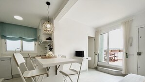 A modern, minimalist room with a white table, chairs, and a bed. There is a window with curtains, a ceiling light, and a view of the outdoors.