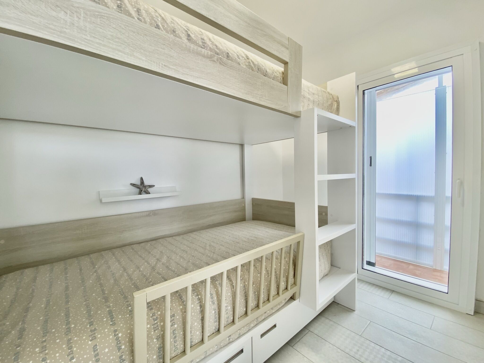 Habitación moderna con literas, estanterías empotradas y puerta corrediza de vidrio que da a un balcón.