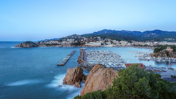 A coastal marina with boats, a pier, and a mountainous backdrop.