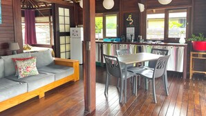 A cozy room with a wooden ceiling, a sofa with floral cushions, a dining table with chairs, a kitchen area with a refrigerator, and a window with curtains. - Heipoe Lodge (Avera, Raiatea)