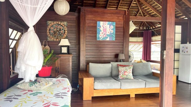 A cozy room with a bed, a sofa, and a dining table. There are decorative pillows and a potted plant. - Heipoe Lodge (Avera, Raiatea)