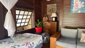 A room with a bed, a sofa, a window with a view, a fan, a potted plant, and a wall with a floral painting. - Heipoe Lodge (Avera, Raiatea)