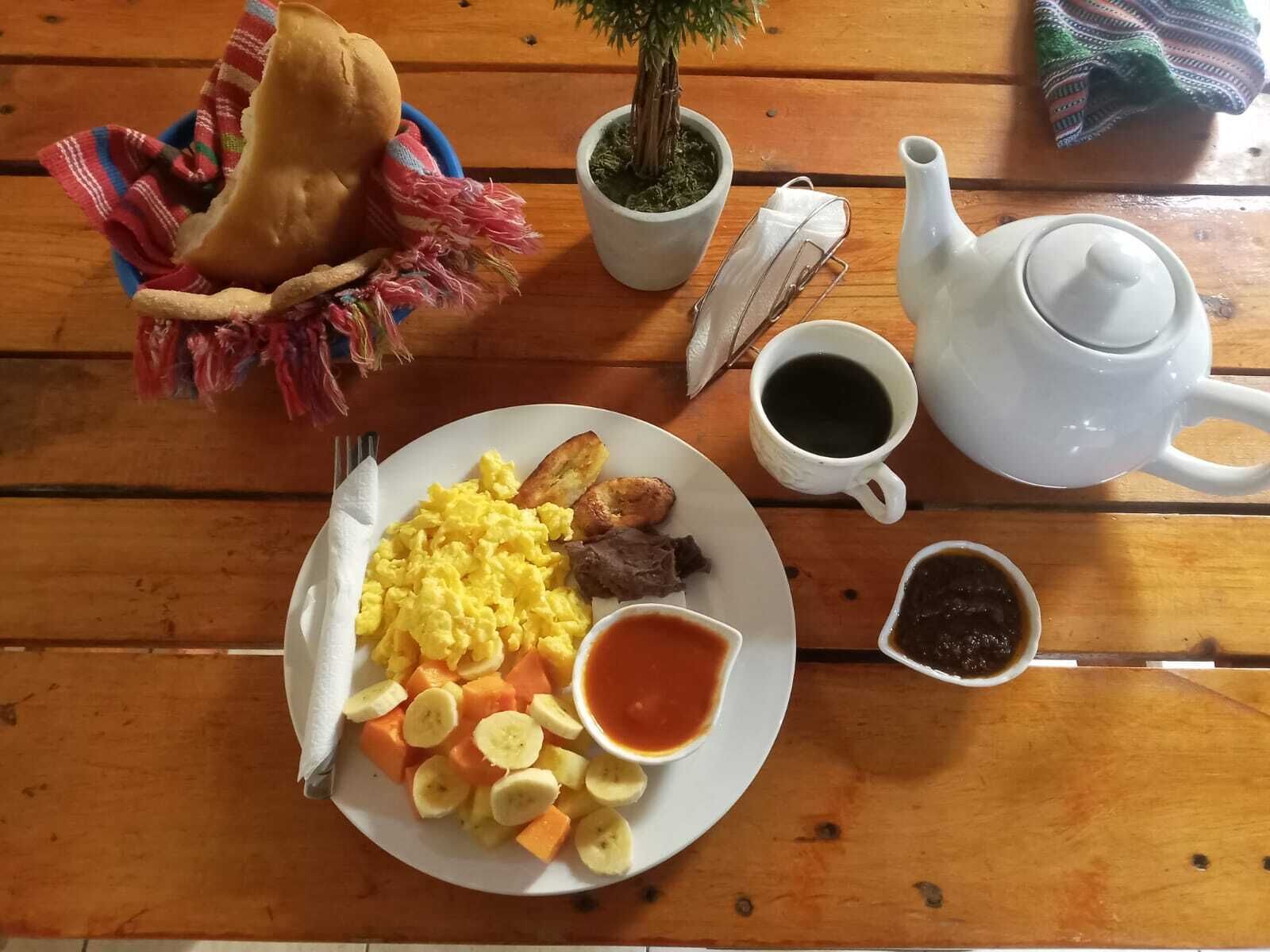 Una mesa de madera con una fuente de huevos revueltos, tostadas y fruta, una tetera, una taza de café y un recipiente pequeño de mermelada.