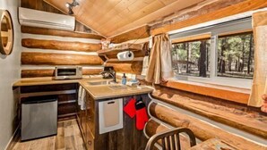 A cabin interior with a kitchen area featuring a sink, a microwave, and a refrigerator. There is a window with curtains, and the walls are made of wooden logs.