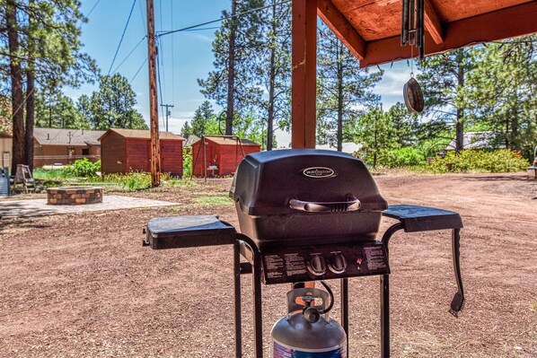 A portable gas grill with a control panel and a propane tank, set up outdoors with a wooden structure overhead. - Spring Valley Retreat | 5 Historic Cabins | Sleeps 24 | Near Grand Canyon & Flagstaff (Bellemont)