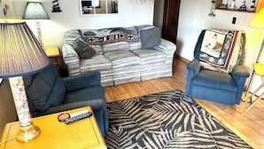 Una sala de estar con un sofá, dos sillones, una mesa de centro, una lámpara, un ventilador de techo, una estantería y una alfombra.