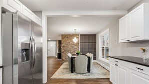 Una cocina moderna con armarios blancos, un refrigerador de acero inoxidable, una zona de comedor con mesa y sillas, y una pared de ladrillo decorativa.