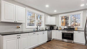 Una cocina moderna con armarios blancos, encimera negra, electrodomésticos de acero inoxidable y grandes ventanales con vistas a árboles.