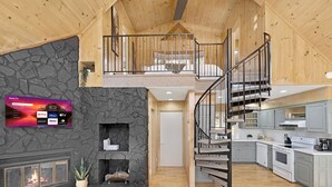 Un interior amplio con una chimenea de piedra, un televisor montado en la pared, una zona de cocina y una escalera de madera que lleva a un nivel superior.