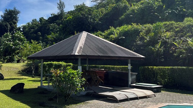 A covered outdoor seating area with a pool, surrounded by lush greenery and a clear sky.