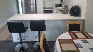 A compact kitchen with white cabinets, a stainless steel refrigerator, a microwave, and a coffee machine. There are two black bar stools and a table with a patterned table runner. -  Bribie Island Dreams (Bongaree)