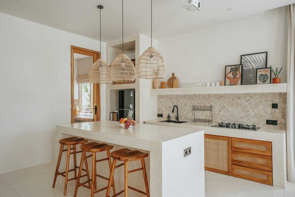 A modern kitchen with a central island, wooden stools, hanging wicker lights, and a tiled backsplash.