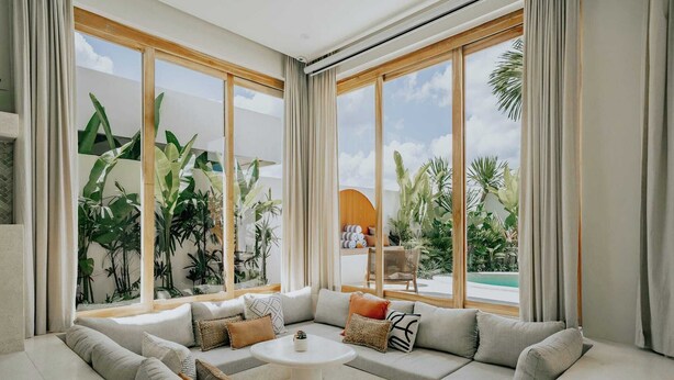 Ein modernes Wohnzimmer mit einer großen Eckcouch, einem kleinen runden Tisch und großen Fenstern mit Blick auf einen Pool und tropische Pflanzen.