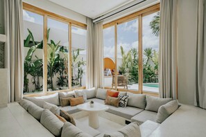 Un salon moderne doté d’un grand canapé d’angle, d’une petite table ronde et de grandes fenêtres donnant sur une piscine et une végétation tropicale.