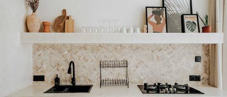 A kitchen with a white countertop, a sink, a gas stove, a backsplash with a herringbone pattern, and a shelf with various items including a vase, a cutting board, and framed pictures.