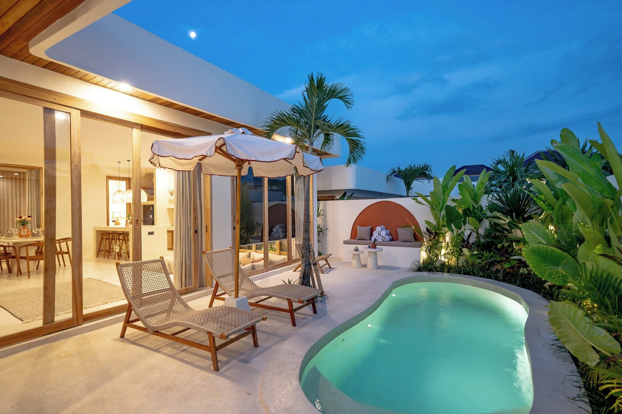 A modern outdoor pool area with lounge chairs and umbrellas, surrounded by lush greenery and a well-lit interior space.