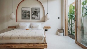 Ein Schlafzimmer mit einem großen Bett, zwei an der Wand befestigten Bilderrahmen, zwei hängenden Pendelleuchten, einem Nachttisch mit Lampe und einem Fenster mit Blick auf Grünflächen.