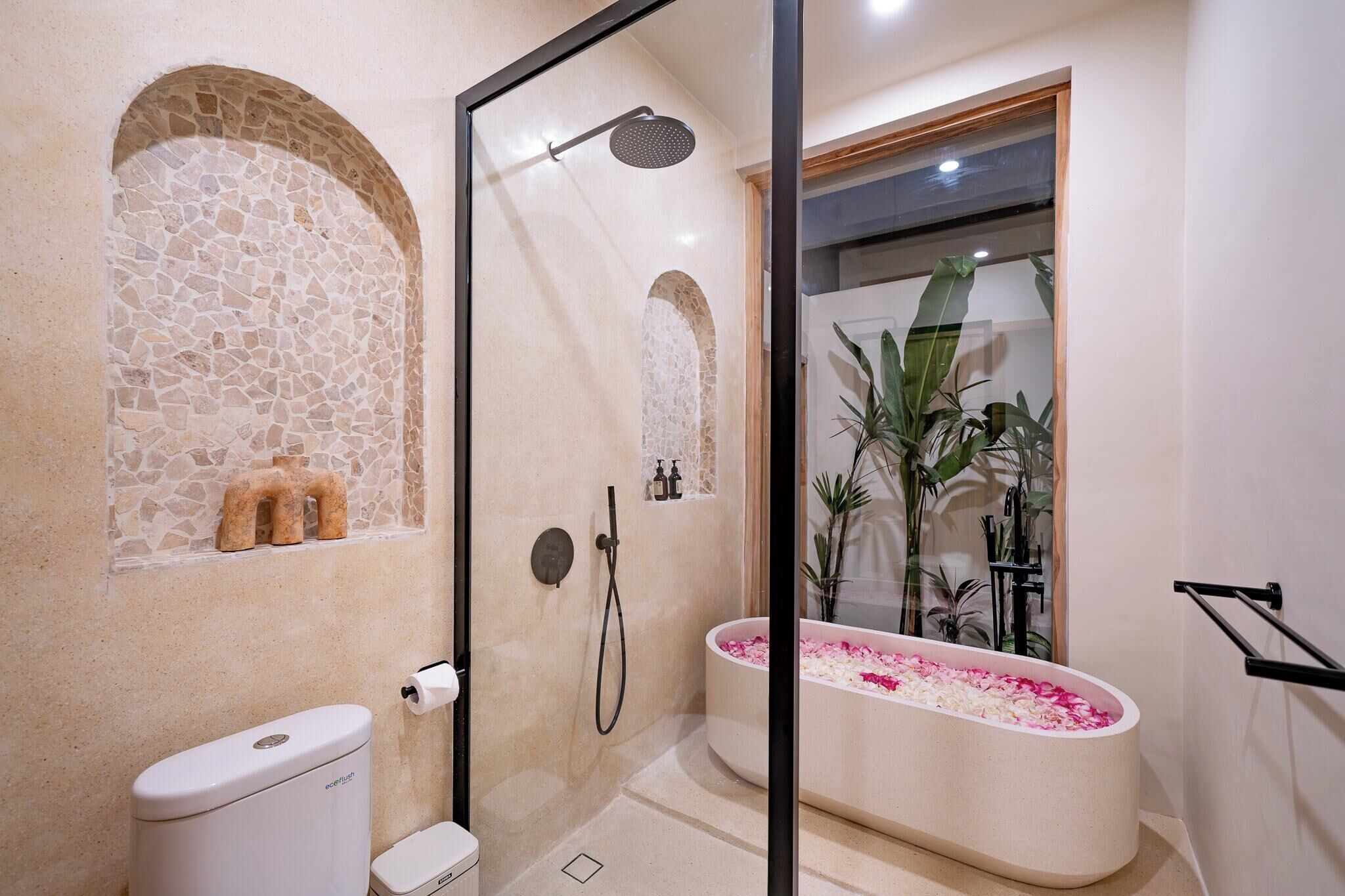 Ein modernes Badezimmer mit einer gläsernen Duschkabine, einer freistehenden Toilette, einer Badewanne mit Rosenblättern und einer Wand aus Steinmosaik.