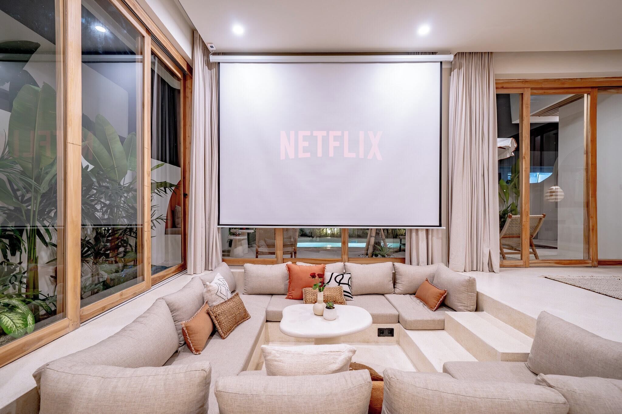 A modern living room with a large flat-screen TV displaying "NETFLIX," a beige sectional sofa, a coffee table, and a view of a pool through large windows.