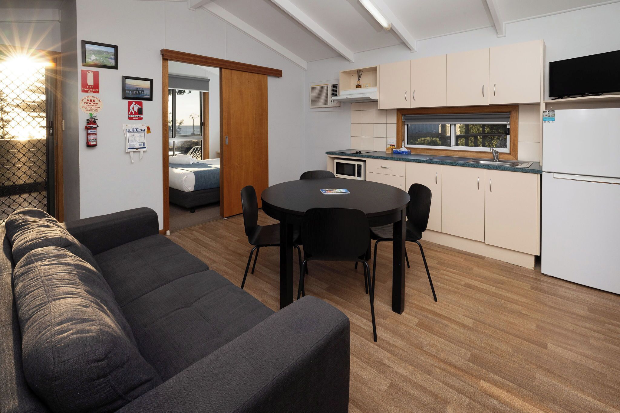 A modern living room with a sofa, dining table, and chairs, a kitchenette with cabinets and a microwave, and a sliding door leading to another room.