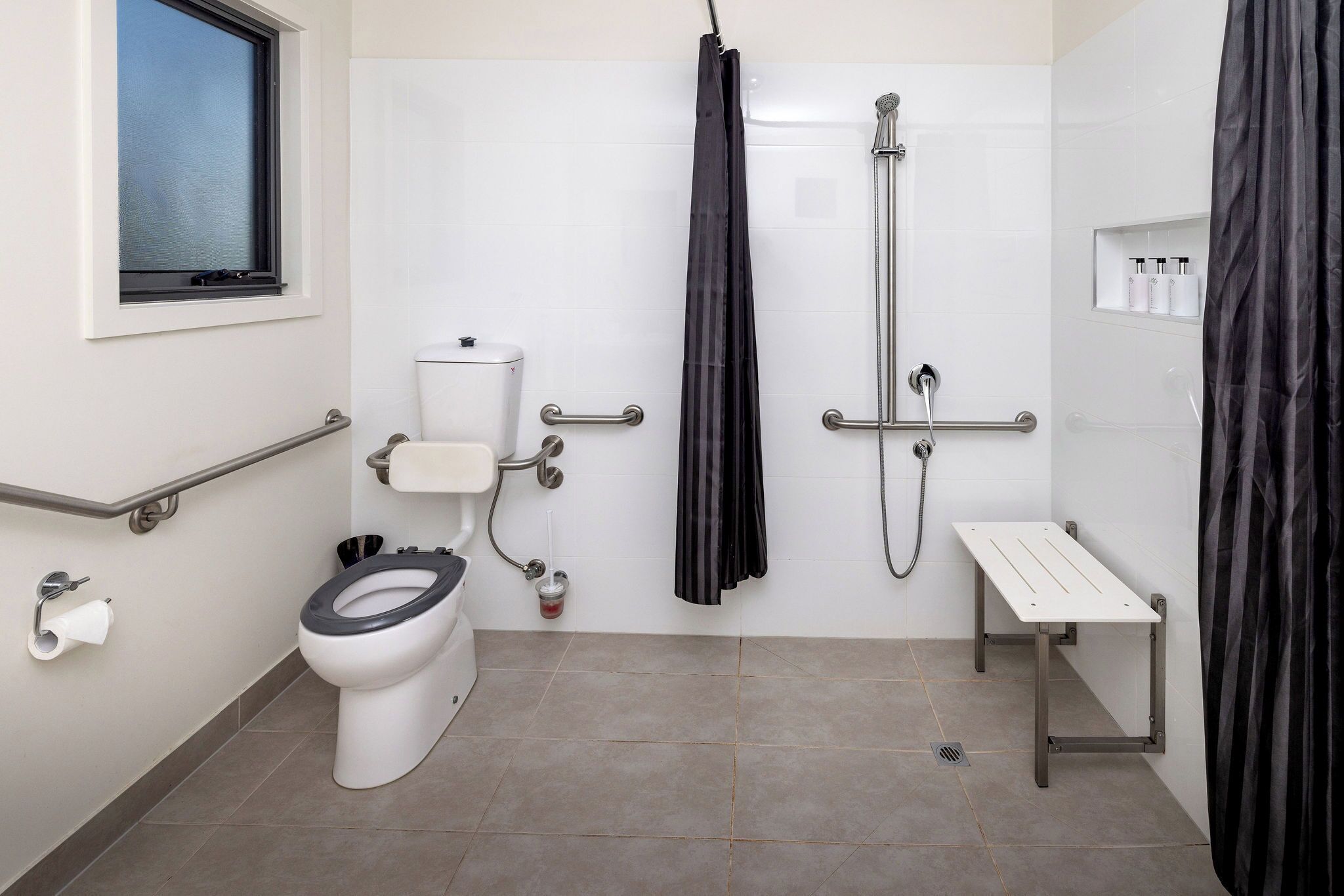 Ein Badezimmer mit Toilette, Dusche, Haltegriff, einer kleinen Sitzbank und einem Fenster.