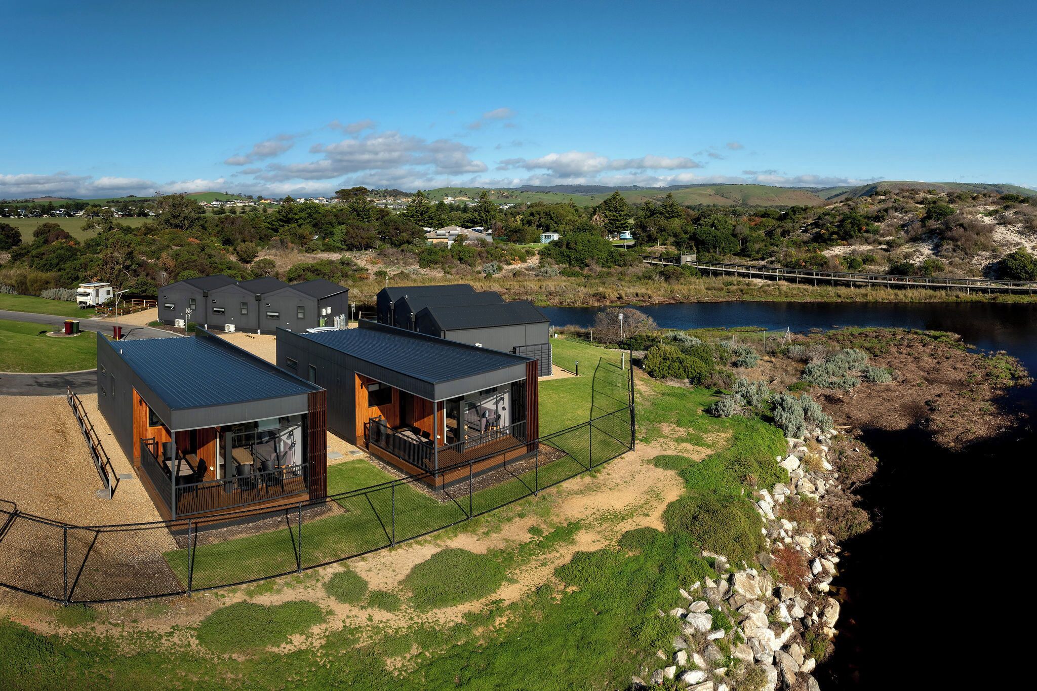 Une maison moderne dotée d’une grande terrasse, d’une cour clôturée et offrant une vue sur un lac et un pont.