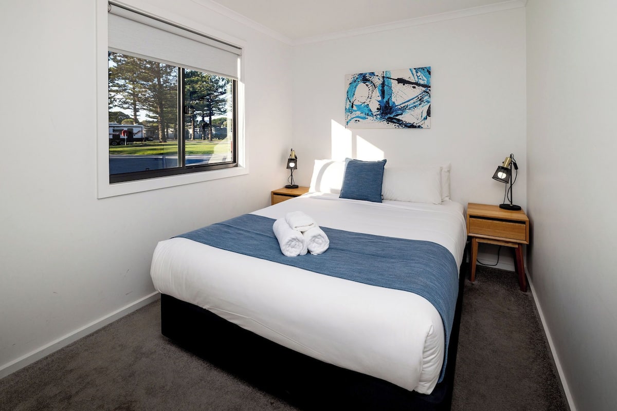 A hotel room with a large bed, a window with a view of trees and a road, a painting on the wall, and two bedside lamps.
