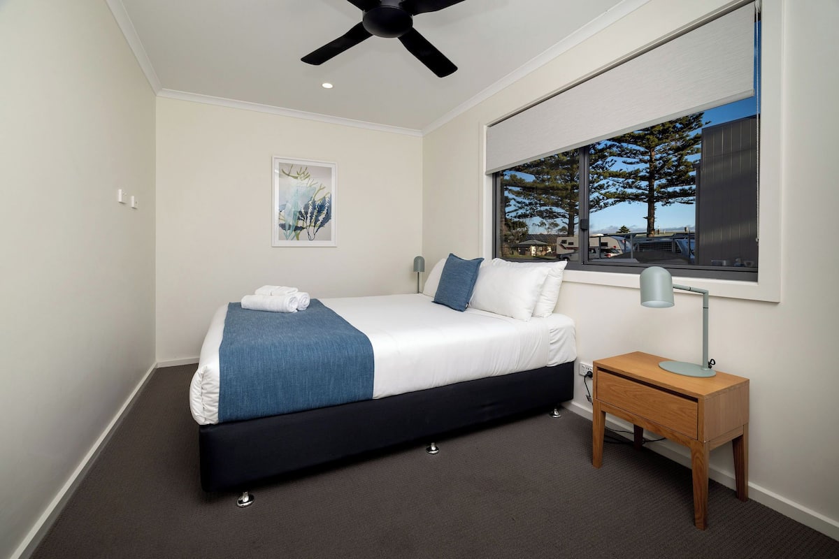A bedroom with a bed, bedside table, lamp, and a window with a view.
