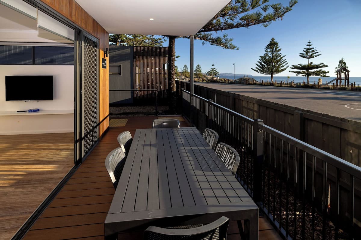 Un área de comedor al aire libre, moderna, con terraza de madera, barandilla de metal y vista al mar.