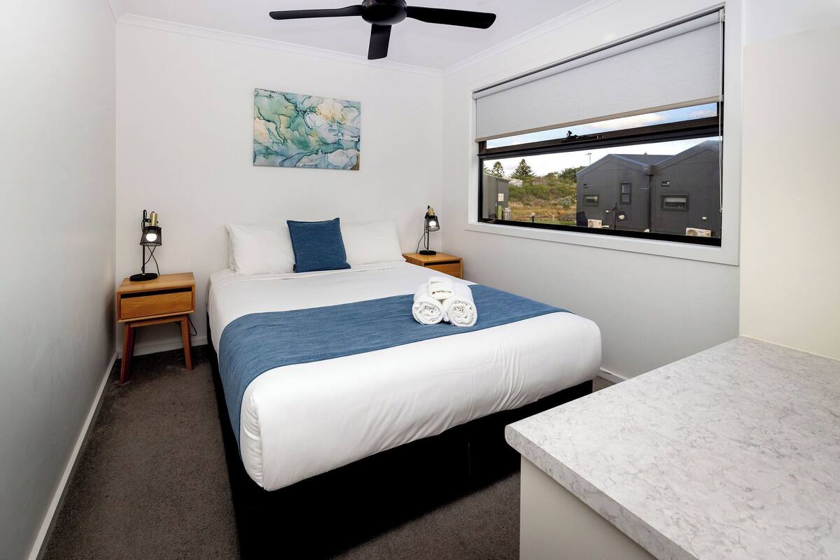 A bedroom with a bed, a ceiling fan, a window with a view, a nightstand, and a painting on the wall.