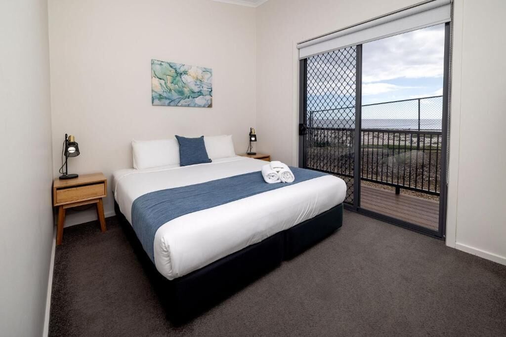 A bedroom with a bed, a nightstand, a lamp, a painting, and a balcony with a view.