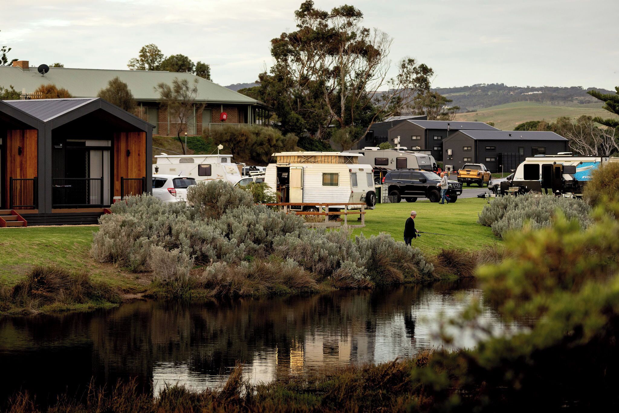 Parque para caravanas con vehículos recreativos estacionados en un área verde, un pequeño edificio con techo inclinado y una masa de agua que refleja el entorno.