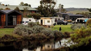 Parque para caravanas con vehículos recreativos estacionados en un área verde, un pequeño edificio con techo inclinado y una masa de agua que refleja el entorno.