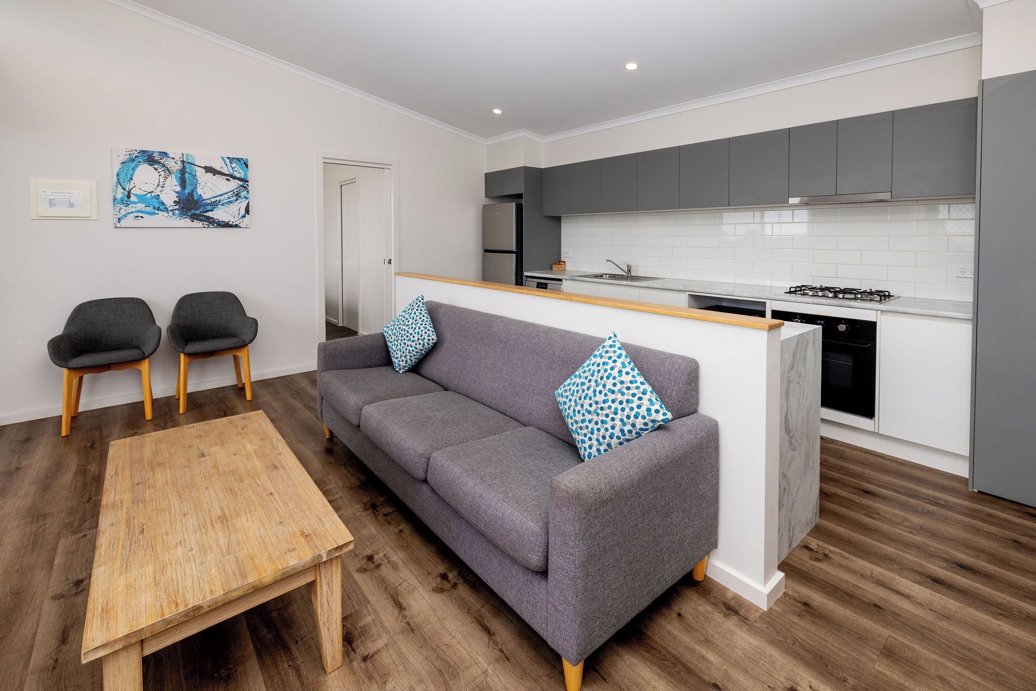 A modern living room with a grey sofa, two armchairs, a wooden coffee table, and a kitchen with grey cabinets and white tiles.