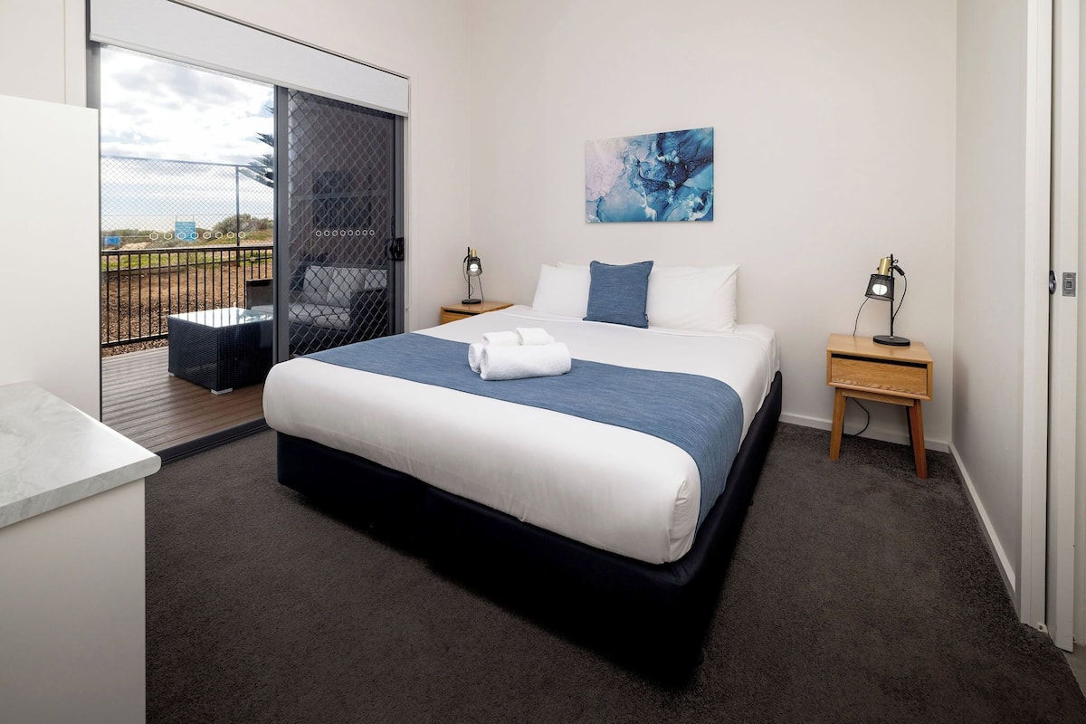 A hotel room with a bed, a desk, a chair, a small table, a nightstand, a painting, and a balcony with a view.
