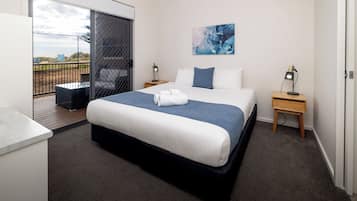 A hotel room with a bed, a desk, a chair, a small table, a nightstand, a painting, and a balcony with a view.