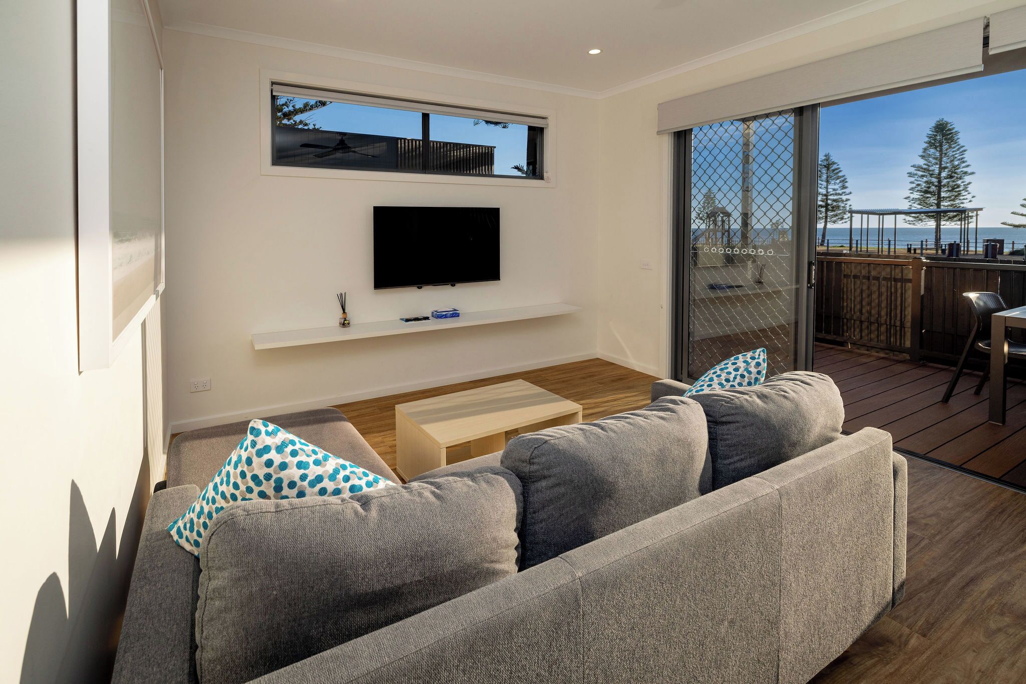 A modern living room with a flat-screen TV, grey sofa, wooden coffee table, and a view of the outdoors through a sliding glass door.