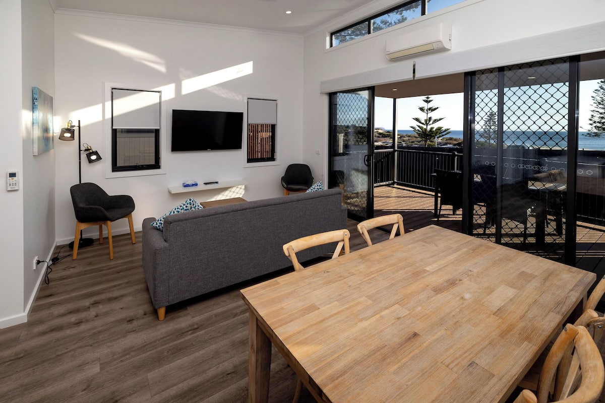 Una sala de estar moderna con una mesa de comedor de madera, un sofá gris y un televisor montado en la pared.