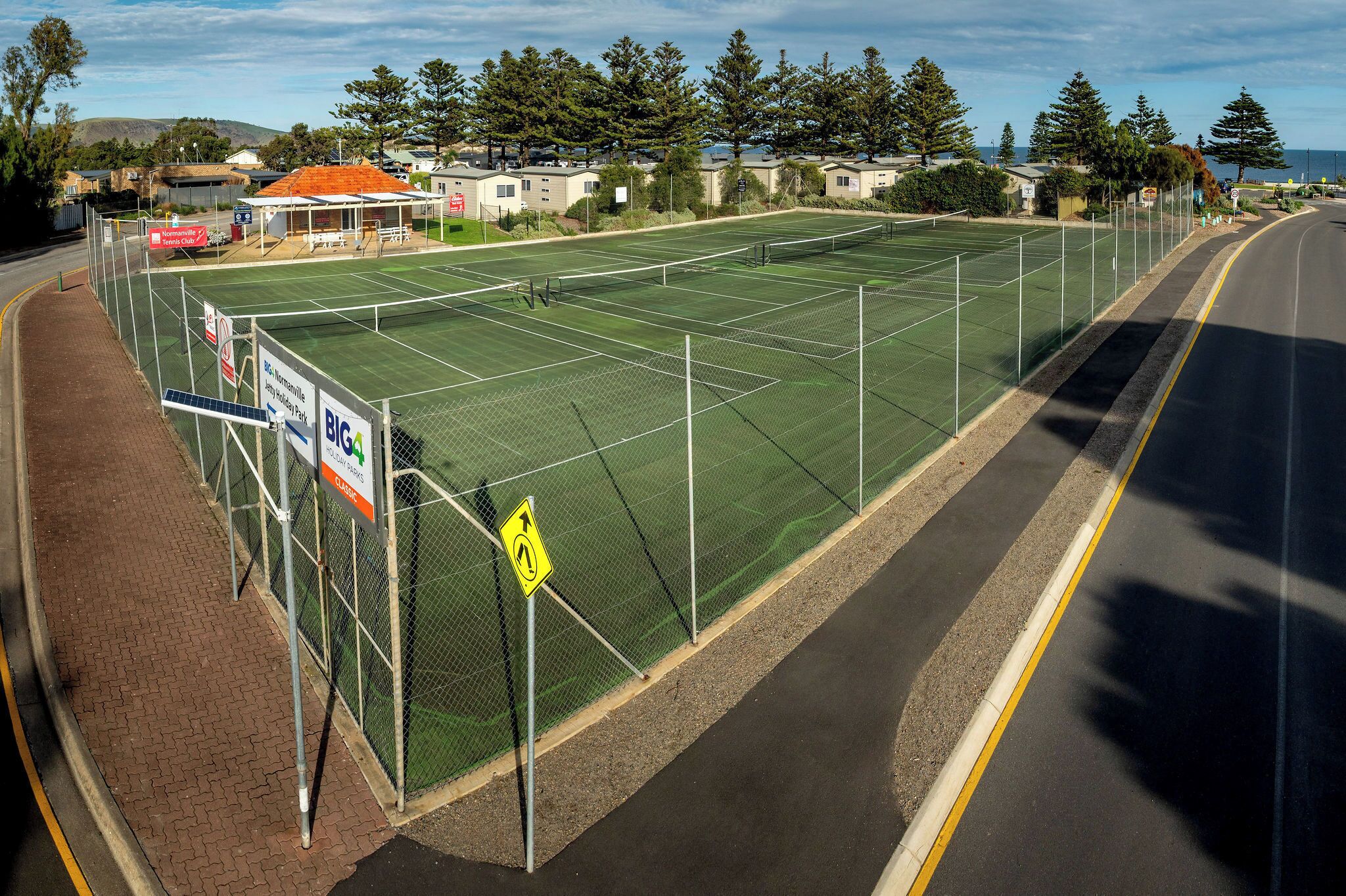 A fenced tennis court adjacent to a road with a pedestrian path.