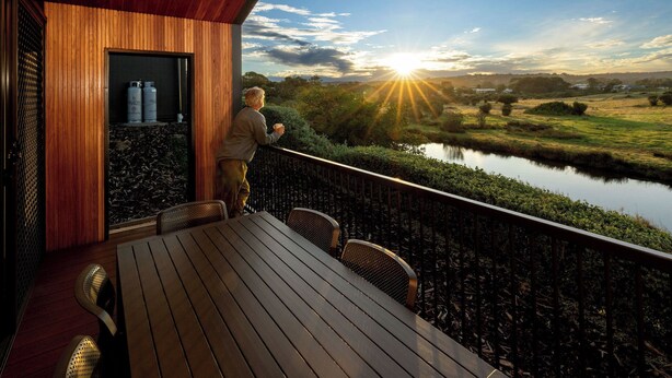 Una terraza de madera con mesa y sillas, vista a un río y un atardecer de fondo.