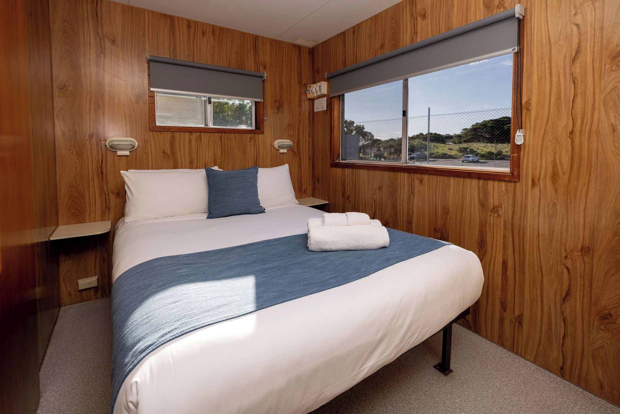 A bedroom with a wooden wall panel, a bed with white and blue bedding, two bedside tables, and a window with a view of the outside.