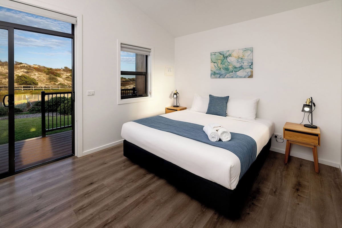 A bedroom with a large bed, a window, a painting, and a view of a grassy area.