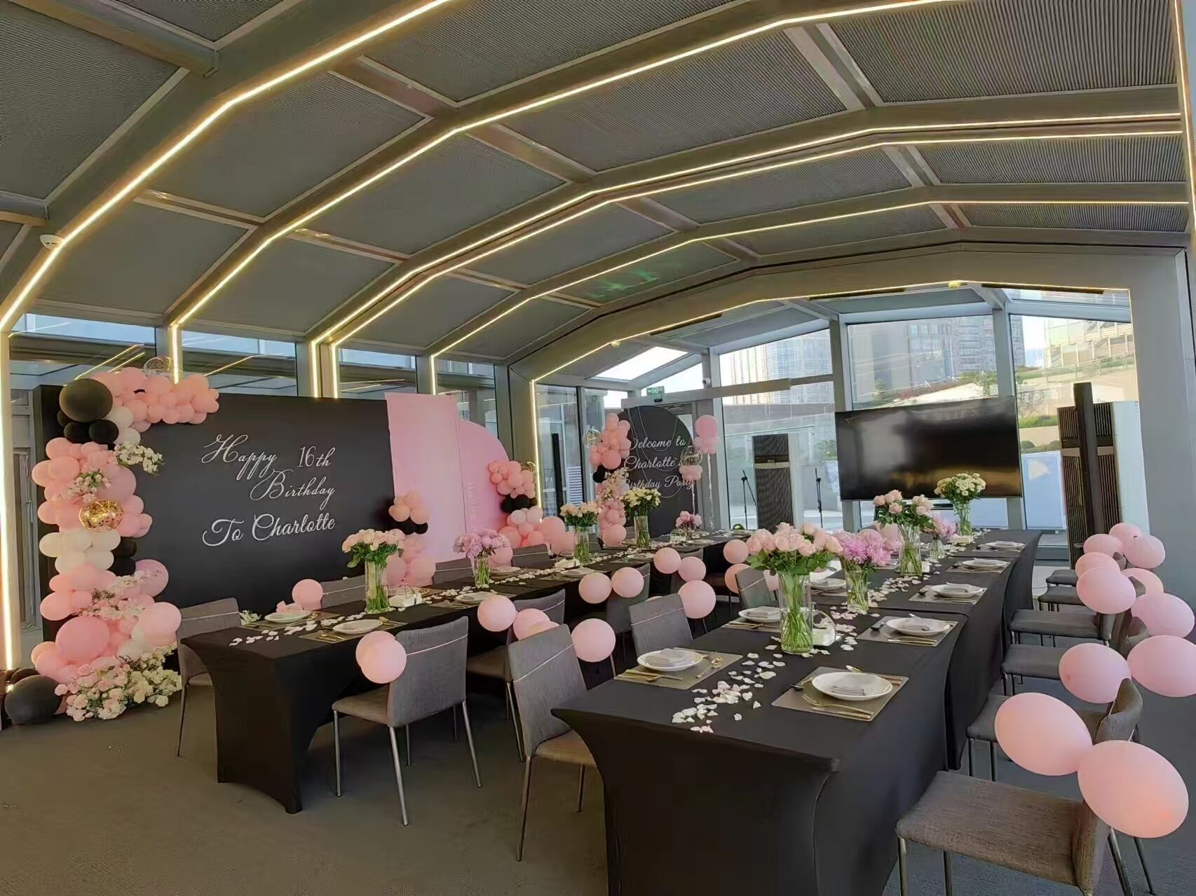 Un salón de banquetes con mesas preparadas para una fiesta de cumpleaños, con un fondo negro que muestra la leyenda 'Happy 16th Birthday To Charlotte'.