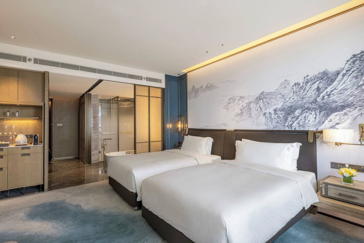 Een hotelkamer met twee bedden, een groot muurschilderij, een bureau met lamp en een zichtbare badkamer door een geopende deur.